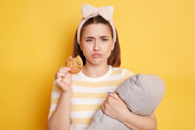 Horizontaal schot van jonge volwassen vrouw met donker haar en haarband die gestreept t-shirt draagt dat koekje eet dat zich voordeed op een gele achtergrond die kussen omhelst terwijl ze junk-ontbijt heeft