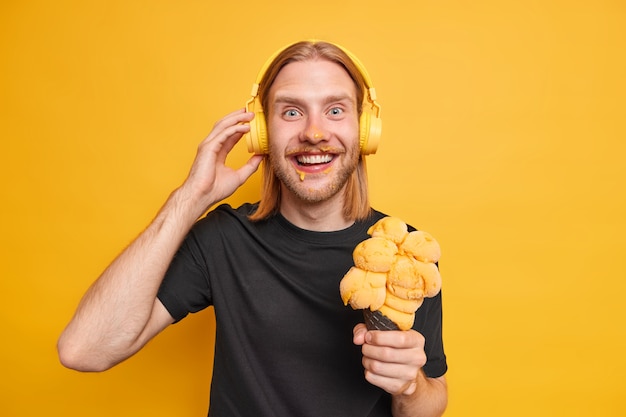 Horizontaal schot van knappe vrolijke man met lang rood haar baard besmeurd gezicht tijdens het eten van heerlijk ijs hoort favoriete audiotrack op radio draagt draadloze koptelefoon gele muur