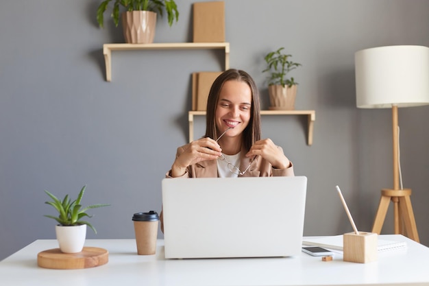 Horizontaal schot van lachende vrolijke vrouw met bruin haar in een beige jas die online op laptop werkt en een zakelijke bijeenkomst heeft die haar bril in handen houdt en geluk uitdrukt