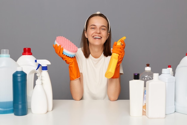 Horizontaal schot van lachende vrouw draagt hoofdband en casual t-shirt met verschillende wasmiddelen om haar huis op te ruimen en huishoudelijke klusjes te doen geïsoleerd over grijze achtergrond