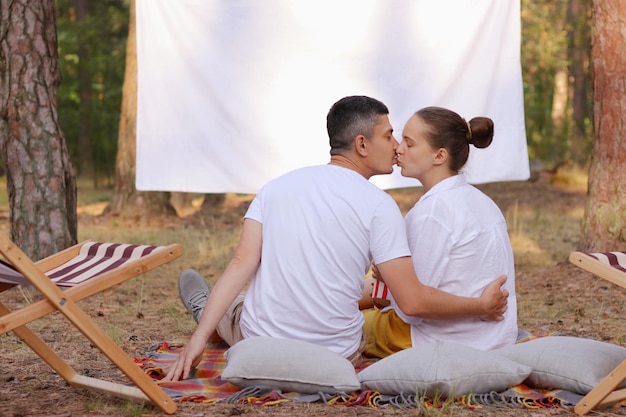 Horizontaal schot van liefdevol romantisch koppel zittend in het bos met overheadprojector met leeg scherm voor promotionele tekst vrouw en man knuffelen en zoenen hun verjaardag vieren