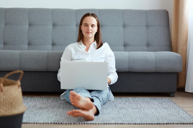 Horizontaal schot van rustige donkerharige jonge volwassen vrouw in wit overhemd en spijkerbroek zittend op de vloer en werkend op laptop, wegkijkend, denkend aan haar werk.