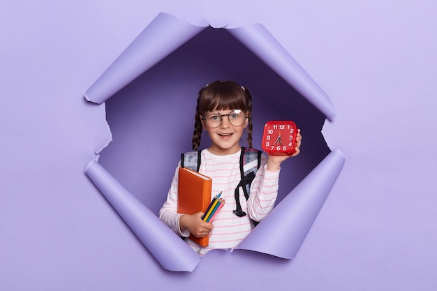 Horizontaal schot van schattig klein meisje in een gestreept shirt met boeken en potloden met wekker die zegt dat het tijd is om terug naar school te gaan poseren geïsoleerd over violette achtergrond