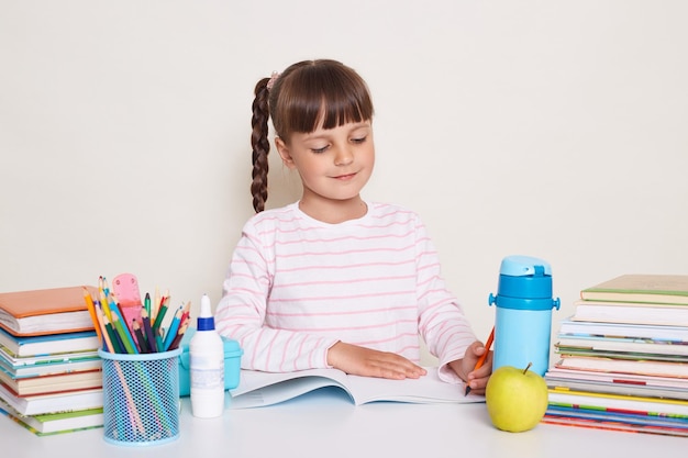 Horizontaal schot van schattig klein schoolmeisje met donker haar en vlechten aan tafel zitten en schrijven in oefenboek poseren omringd met boeken die geconcentreerd en attent glimlachen