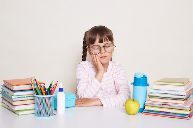 Horizontaal schot van slaperig klein schoolmeisje met donker haar en vlechten zittend aan een bureau omringd met boeken geïsoleerd op een witte achtergrond houdt de ogen gesloten