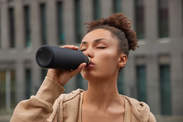 Horizontaal schot van sportieve vrouw drinkt verfrissend water uit fles houdt ogen gesloten voelt dorstig na training draagt sweatshirt leidt gezonde levensstijl Vermoeide atletische vrouwelijke oefeningen regelmatig