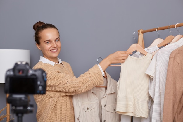 Horizontaal schot van tevreden vrouwelijke blogger met knotkapsel die video opneemt met behulp van camera met statief, pratend over modieuze outfits in haar vlog met kleding op hang op plank