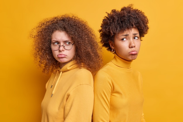 Foto horizontaal schot van twee ongelukkige, diverse vrouwen die tegenover elkaar staan en niet spreken na ruzie portemonneelippen gekleed in vrijetijdskleding geïsoleerd over gele muur. negatieve emoties concept