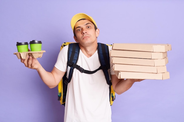 Horizontaal schot van verbaasde man in gele pet en t-shirt met pizza en papieren kopjes koffie geïsoleerd op lila achtergrond schouderophalend en kijkend naar camera