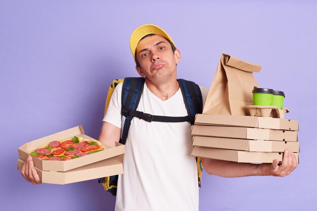 Horizontaal schot van verwarde onzekere koerier houdt pizzadozen en afhaalkoffie in de hand geïsoleerd op paarse achtergrond schouderophalend kent het adres van de klant niet