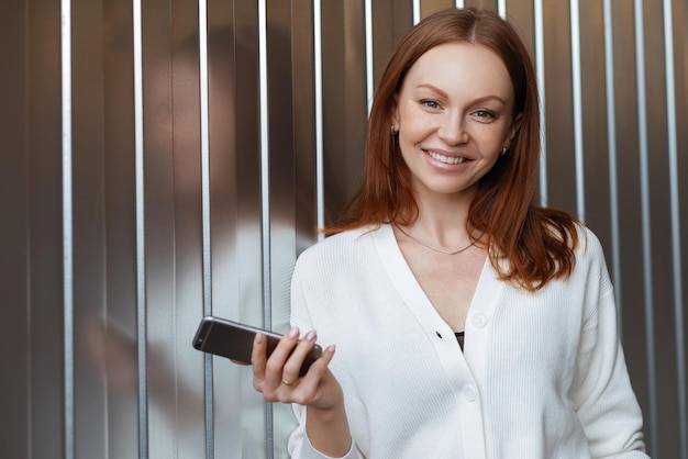 Horizontaal schot van vrolijke vrouw met geverfd haar brede glimlach chats online via smartphone maakt gebruik van appliaction voor online bankieren draagt elegante witte trui staat over muur Technologie concept