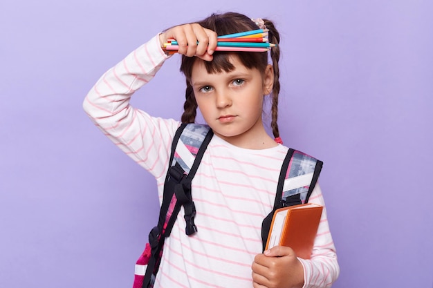 Horizontaal schot van ziek vermoeid meisje in een gestreept shirt met notitieboekje en pennen kijkend naar camera met uitgeputte gezichtsuitdrukking die lijdt aan hoofdpijn poseren geïsoleerd over lila achtergrond