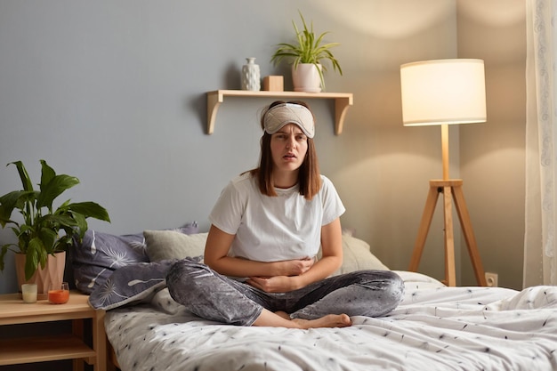 Horizontaal schot van zieke ongezonde trieste vrouw met slaapmasker zittend op bed in knusse slaapkamer met vreselijke sterke buikpijn fronsend gezicht van pijn