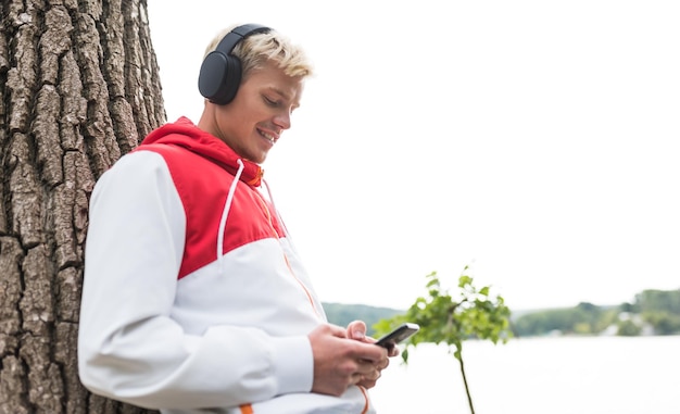 Horizontaal zijschot van een jonge blonde man die geniet van het weer in het stadspark, wachtend op zijn vriendin die lacht terwijl hij naar zijn favoriete nummer luistert met behulp van mobiele telefoon buiten in het park lifestyle concept