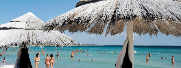 Horizontal banner or header with a beautiful beach resort with straw umbrellas on a blue sky and white clouds On the background bathers in swimsuits Beach life and lifestyle concept