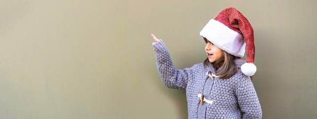 Horizontal banner or header with happy confident little girl wearing santa claus hat pointing at green copy space Funny kid playing against green wall outdoors in Christmas time