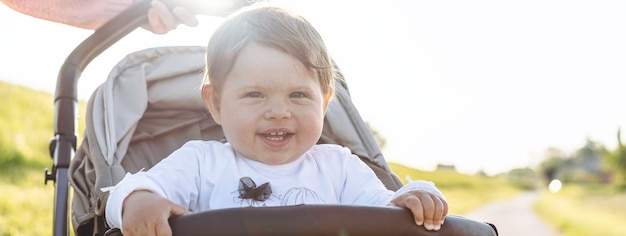 Horizontal banner or header with happy laughing baby girl enjoying a walk in countryside sitting in the modern stroller Sweet child in buggy showing first teeth