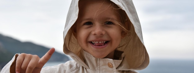 Horizontal banner or header with a little smiling girl wearing a raincoat jacket with hood against the stormy sky She lifts her index finger Childhood Environmental and Climate change concepts