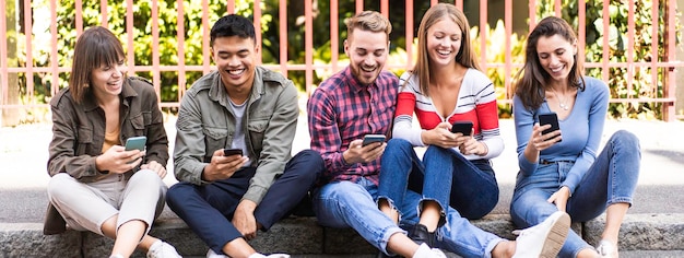 Horizontal banner or header with multicultural friends laughing using smartphone sitting on the sidewalk in the city Young people addicted by mobile smart phones