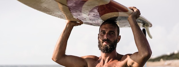 Horizontal banner or header with surfer holding his surfboard on the head Hipster man standing on the beach and waiting big waves for surfing Fit bearded man training with surfboard to sea