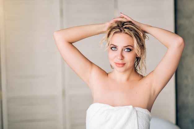 Horizontal closeup portrait of lovely young caucasian blonde woman with beautiful blue eyes and healthy clean skin after spa treatments, covering naked body with towel, looking smiling at camera.