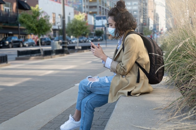 Horizontal frontal shot of seated woman with mask sending\
message with mobile phone and hanging backpack, with copy\
space.