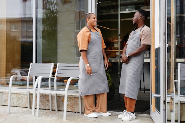 Horizontal full shot of young black man and woman wearing aprons standing at cafe entrance talking a...