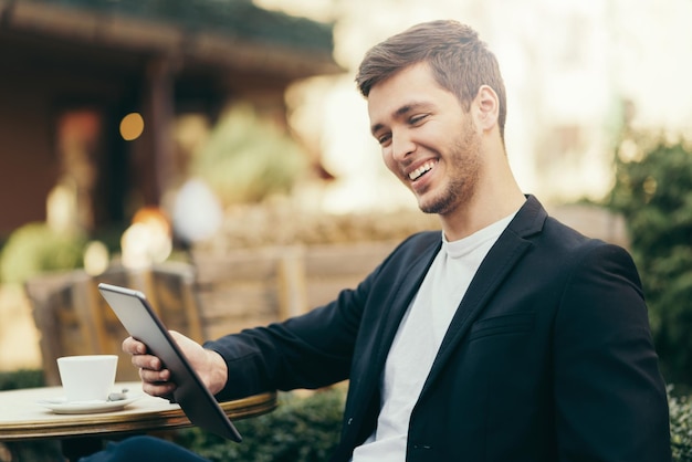 Horizontal happy caucasian man look to the digital tablet sitting in cafe with cup of coffee Business man having breakfast sitting on beautiful terrace with plants Reading and browsing internet