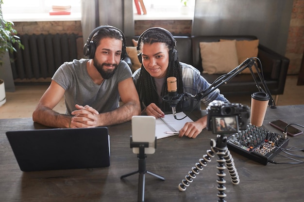 Photo horizontal high angle shot of multiethnic man and woman sitting at desk in loft room looking at smartphone camera