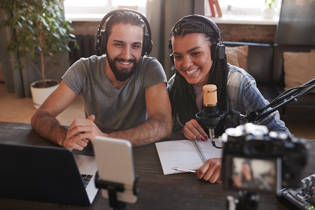 Photo horizontal high angle shot of two happy multiethnic influencers sitting at desk in loft room smiling at smartphone camera