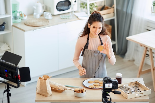 カメラで自家製クッキーを示す彼女のキッチンで一人で立っている若い大人の女性の水平方向のハイアングルショット