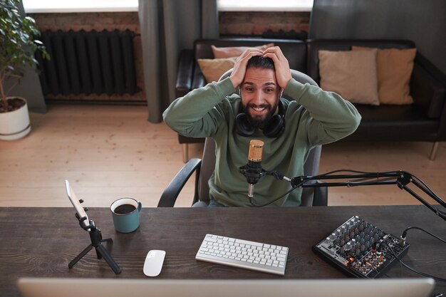Foto inquadratura orizzontale dall'alto di un giovane influencer mediorientale barbuto moderno che indossa abiti casual seduto alla scrivania in una stanza soppalcata eccitandosi