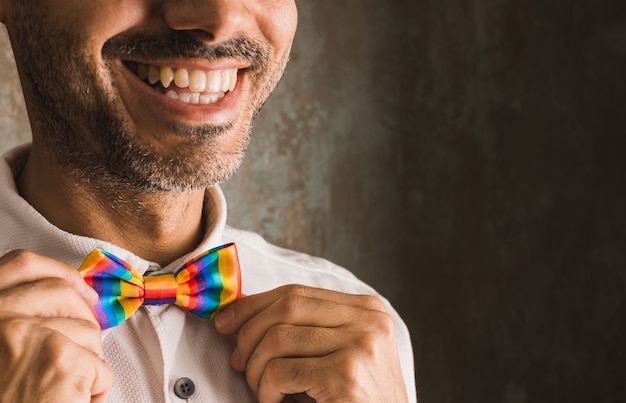 Horizontal image of brunette guy with beard smiling in white\
shirt and lgbtqi + rainbow bow tie on left side of image on worn\
wall illuminated with soft side light