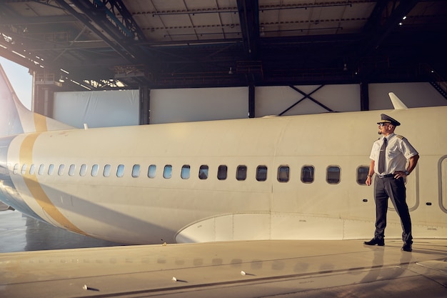 Immagine orizzontale di un bel pilota in uniforme da lavoro in piedi sull'ala dell'aereo passeggeri mentre guarda lontano