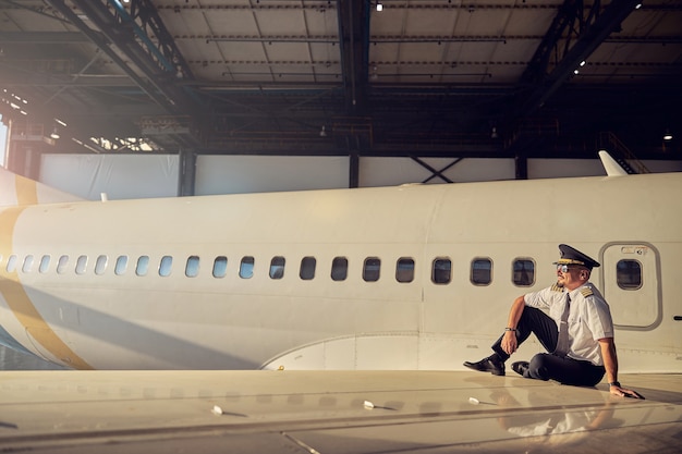 Horizontal image of thoughtful pilot sitting on the airplane and looking to the distance isolated on fuselage background