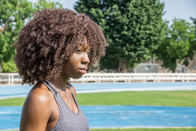 Horizontal image of young black female athlete with afro hair looking to the side with blue running track grass and green trees in background with harsh natural lighting