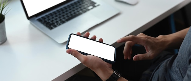 Horizontal image of young man holding smart phone with blank screen while sitting at office desk.