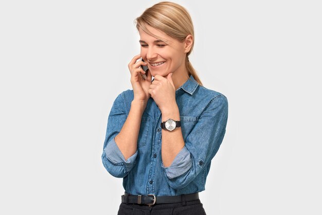 Horizontal indoor image of young pretty blonde woman in denim shirt smiling and talking on smart phone to her friend looking cheerful and happy posing on white studio background Real human emotions