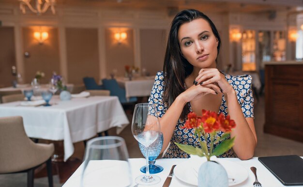 Horizontal indoor portrait of brunette young elegant woman wearing dress sitting in the restaurant looking to the camera Caucasian female posing in cafeteria and waiting her friends Copy space