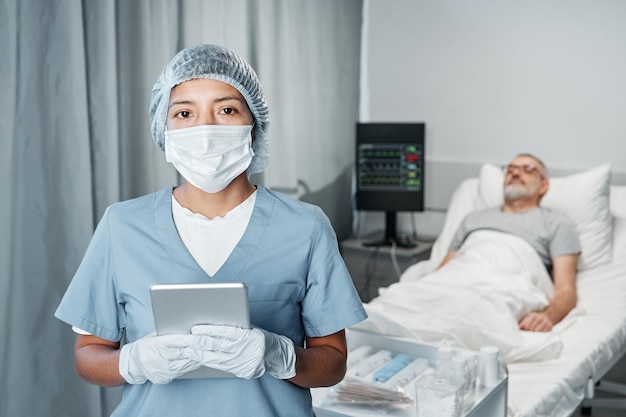 Horizontal medium portrait of unrecognizable medical worker wearing mask holding digital tablet looking at camera her patient lying in bed on background