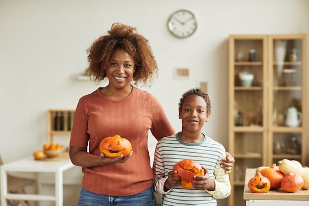 Horizontal medium portrait of young adult woman and her preteen son standing together in living room holding pumpkins they carved smiling at camera