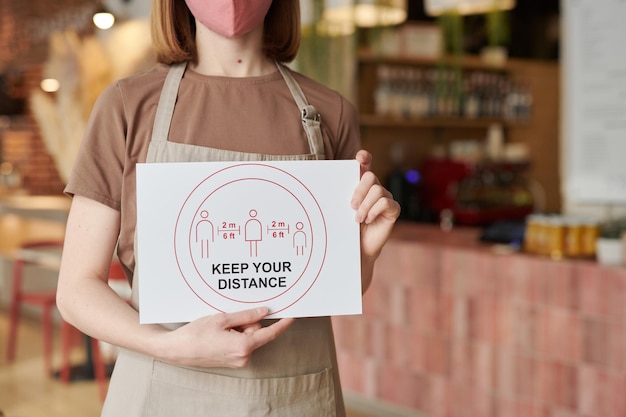 Horizontal medium section shot of unrecognizable waitperson standing in cafe holding Keep Your Distance sign