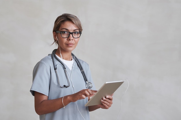 Horizontal medium studio portrait of modern female doctor wearing eyeglasses and earphones using dig