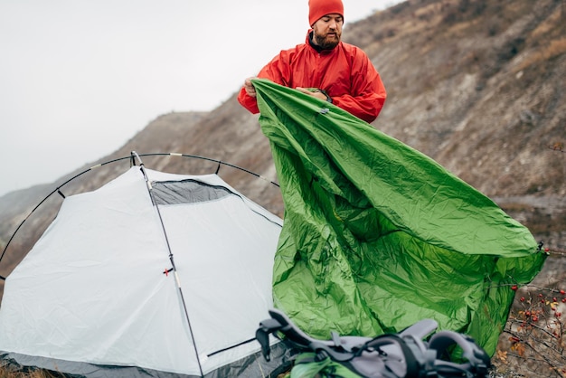 Horizontal outdoors image of authentic young hiker man prepares his tent in mountains Traveler bearded male relaxing after mountaineering Travel people and healthy lifestyle concept