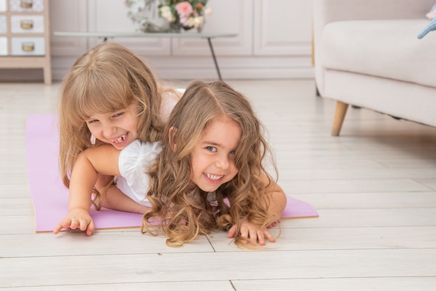 Horizontal photo of couple little girls playing on yoga mat smiling in living room, healthy life habits and lifestyle concept.