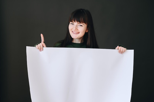 Горизонтальное фото девушки, которая держит в руках белую бумагу и указывает указательным пальцем вверх