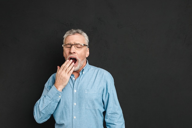 Horizontal photo of mature unshaved man 60s with grey hair wearing eyeglasses being sleepy and yawning because of insomnia, isolated over black wall