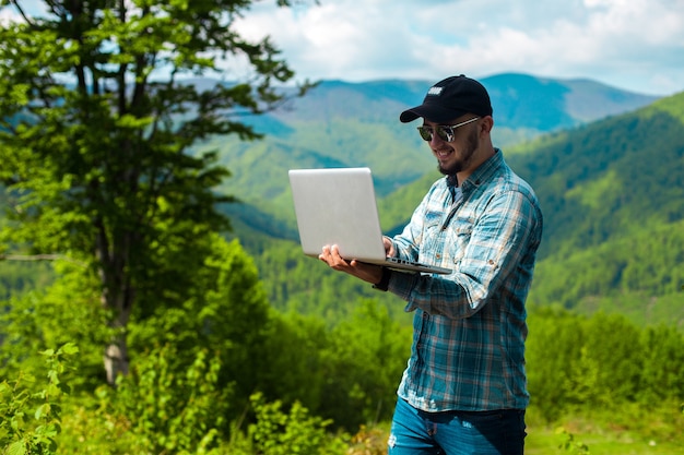Horizontal photo of stylish young boy smiling and working on laptop in the mountains