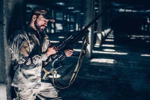 Photo horizontal picture of soldier standing very close to column with his back and recharging riflle. he is in a big hangar alone. guy is taking a short break. he is going to be ready to fight again.