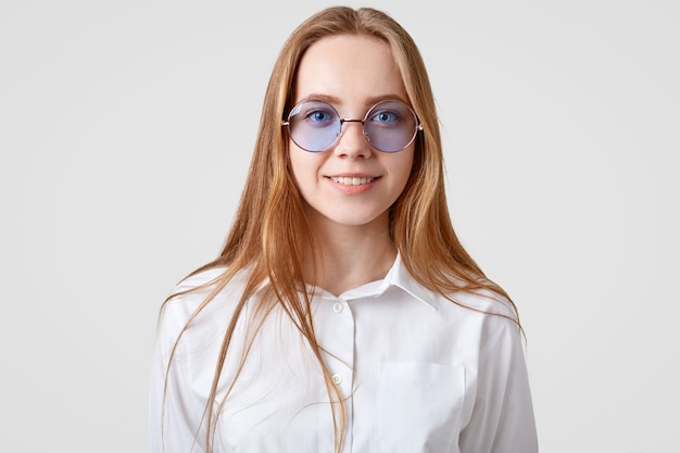 Horizontal portrait of cheerful female student wears white shirt, trendy round shades, looks directly at camera, models in studio alone